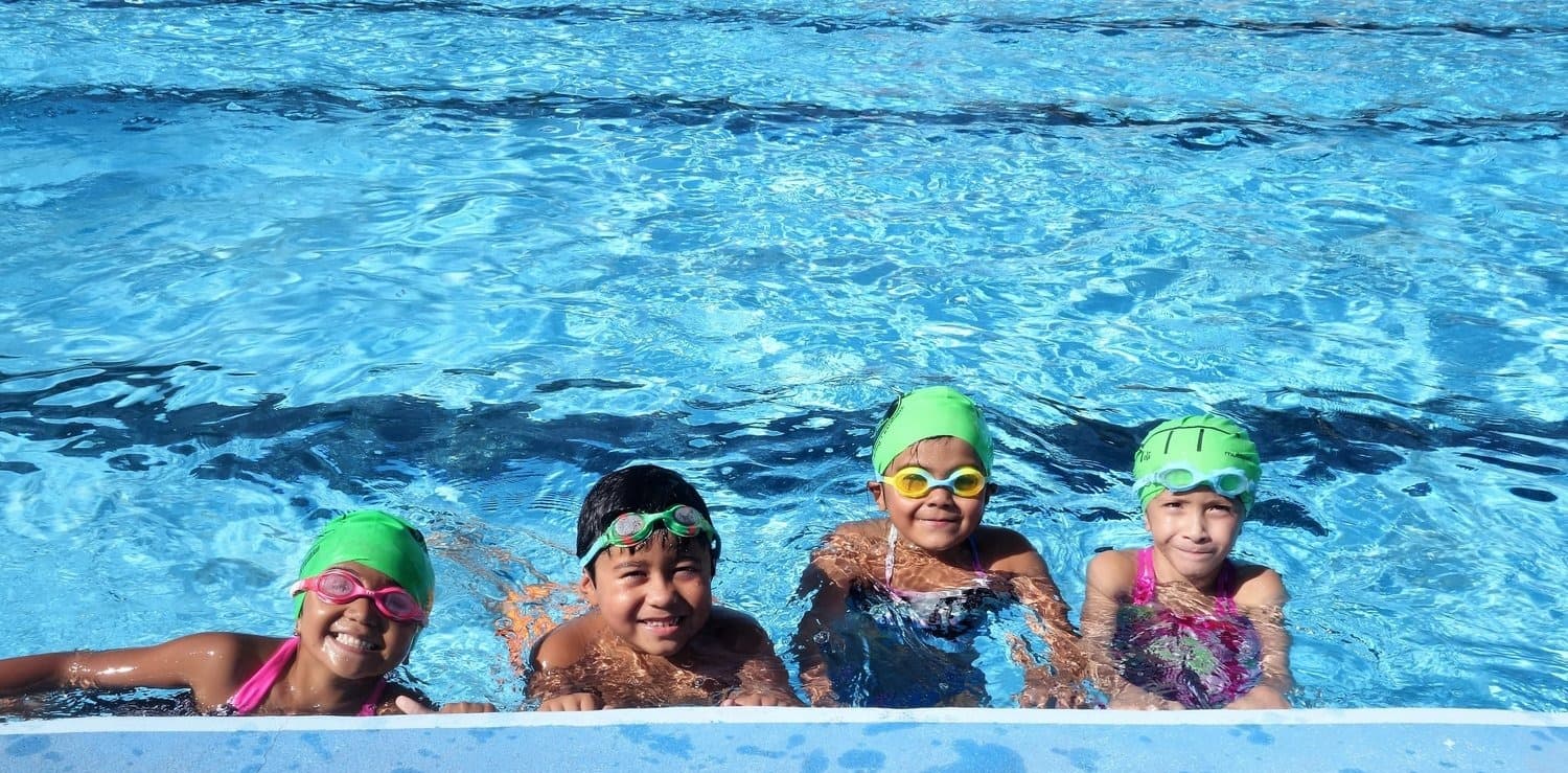 Four smiling kids in goggles and green ViVe swim caps holding the edge of a pool