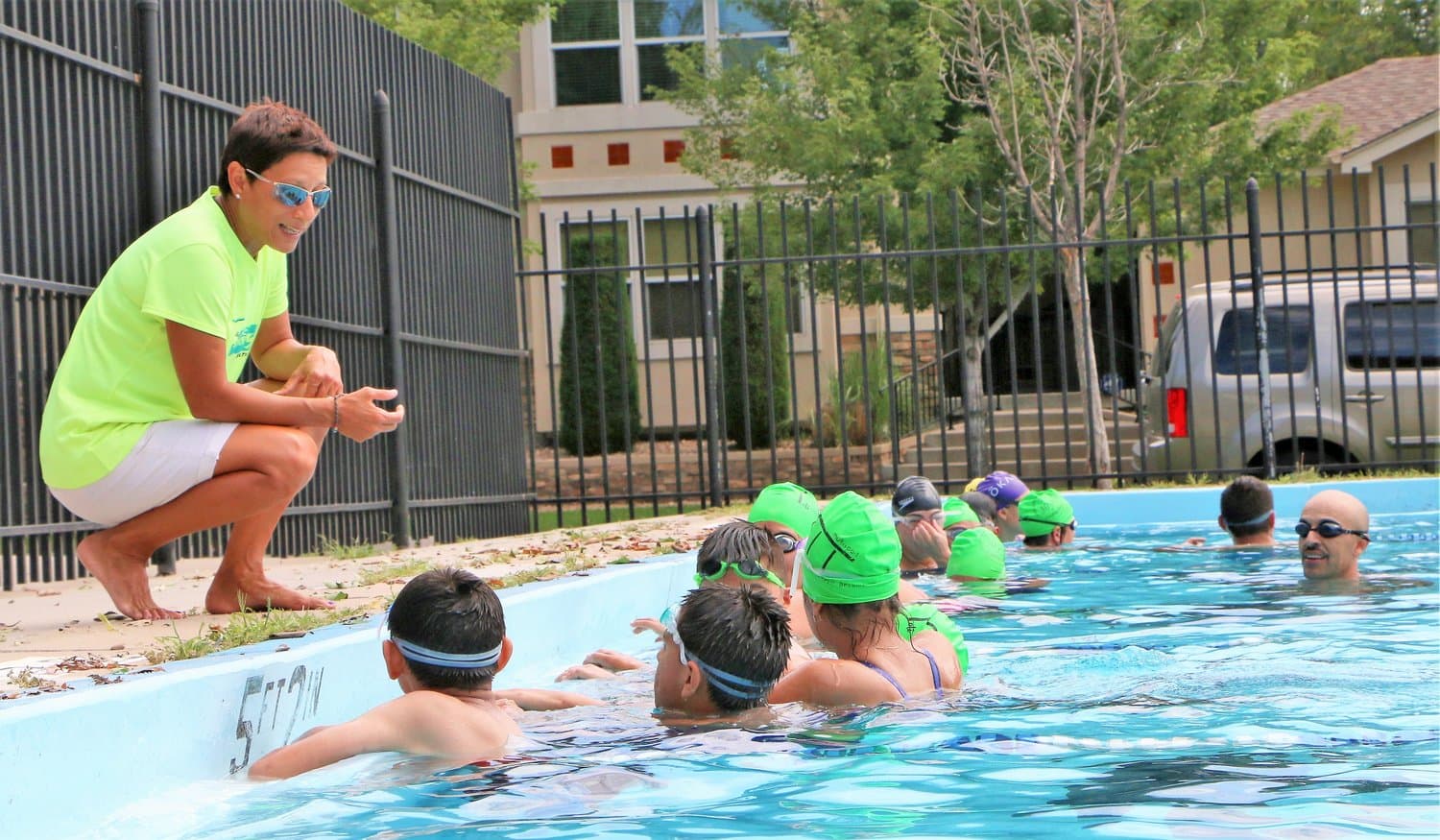 A ViVe coach in a lime shirt crouches at the pool edge, teaching a row of kids in green swim caps