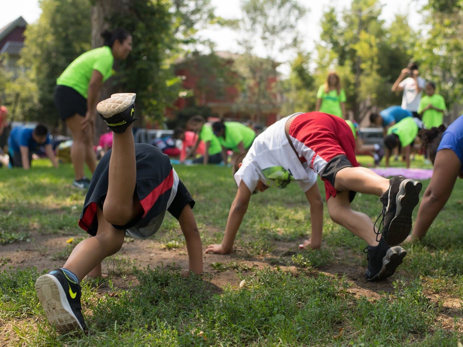 Kids doing a crawling exercise on the grass in a park while coaches in lime ViVe shirts cheer them on